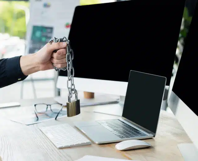 Cropped view of programmer holding padlock on chain near computers with blank screen in office